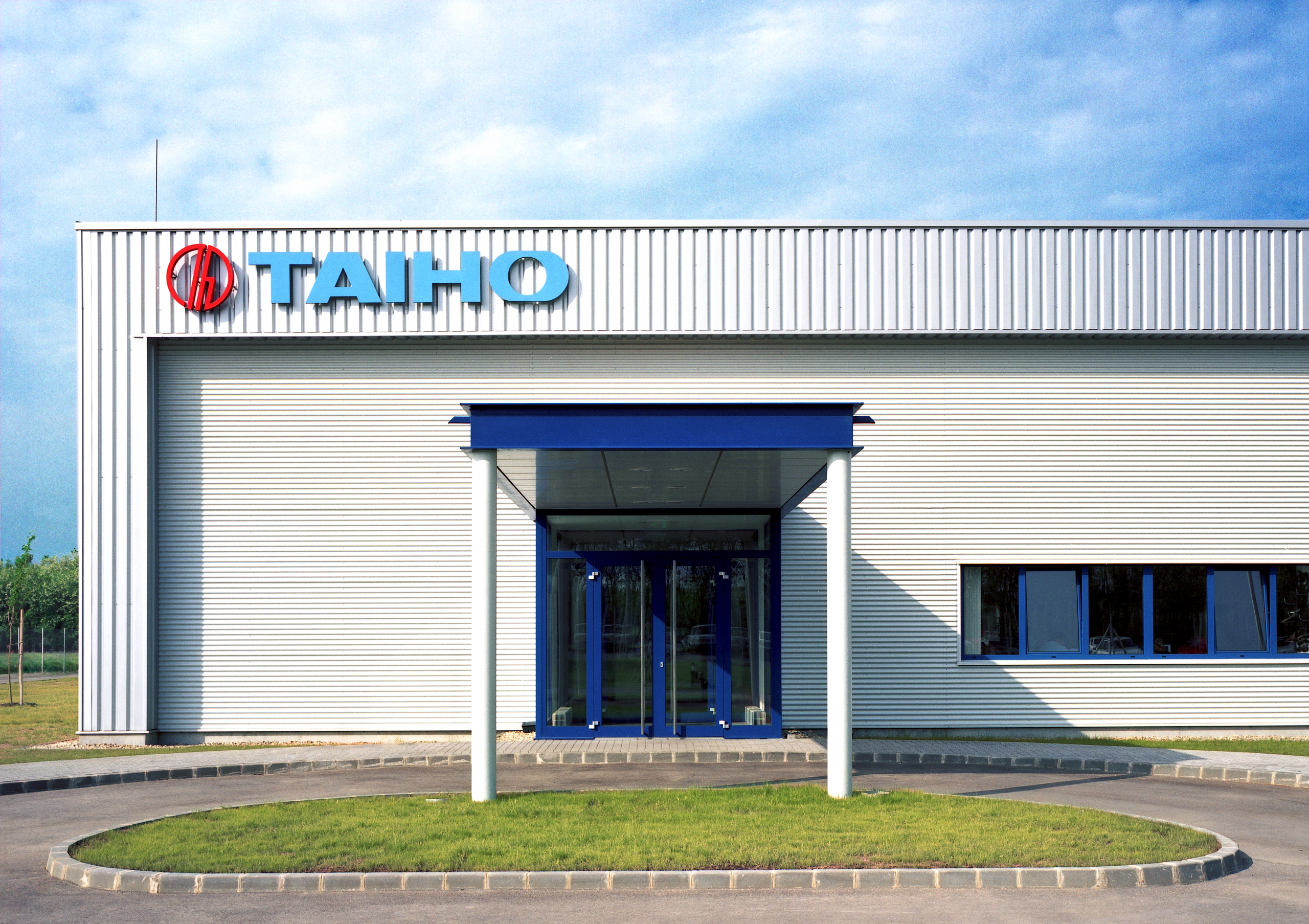 Factory Taiho Corporation of Europe Kft. built by Takenaka Europe in 2005. Modern industrial building facade with vertical white sheet metal panels, large blue entrance portal and company logo "TAIHO" in red and blue.